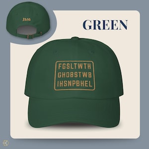 Green baseball cap with the text "GREEN" and "J3:16". The front of the cap features a gold rectangular design with the text "FGSLTWTH GHOBSTWBI HSNPBHEL". The cap has an adjustable strap.