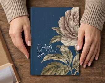 May include: A rectangular prayer journal with a dark blue cover featuring a large, detailed illustration of a beige peony flower and green leaves. The text "Caroline's Prayer Journal" is printed in white script on the left side of the cover.
