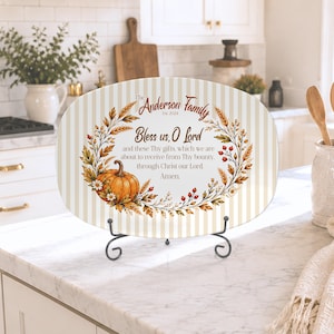 May include: An oval ceramic serving platter featuring a beige and white vertical striped background. It displays a wreath of autumn leaves, berries, and a pumpkin, with the text: The Anderson Family Est. 2024 Bless us, O Lord and these Thy gifts.