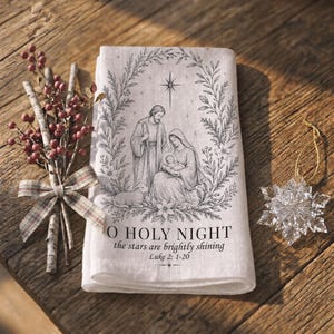 Pode incluir: Um pano de tecido retangular em tom esbranquiçado com uma ilustração de traço preto da cena da Natividade dentro de uma coroa de flores. O texto O HOLY NIGHT, the stars are brightly shining e Luke 2: 1-20 está centrado abaixo da imagem.