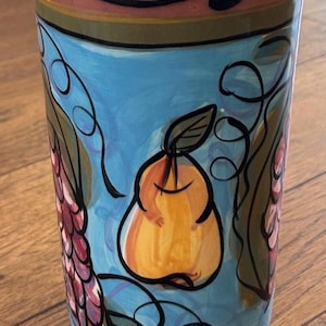 May include: A cylindrical ceramic tumbler featuring a hand-painted light blue background with a central yellow pear illustration. The design includes dark green vine accents, black outlines, and clusters of purple grapes on the sides with a rim border.
