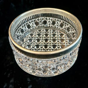 May include: A clear, cut-glass bowl with a silver-colored rim. The bowl features a detailed, geometric pattern throughout its surface. The rim has a decorative design. The bowl is likely for serving or display.