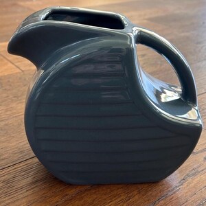 Puede incluir: Una jarra de cerámica gris oscuro con acabado brillante, que presenta un cuerpo redondeado con líneas horizontales en relieve, un pico vertedor ancho y un asa curva que conecta el borde superior con el lado inferior del recipiente.
