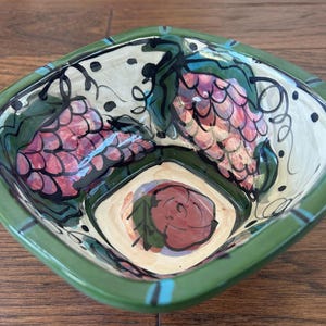 Puede incluir: Un cuenco cuadrado de cerámica con borde verde e interior pintado. El cuenco presenta un diseño de uvas rojas, hojas verdes y una rosa roja. El fondo es de color crema con puntos y remolinos negros.