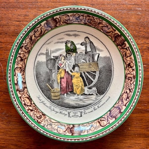May include: A round ceramic plate with a green rim and a brown decorative border featuring a central illustration of people with a cart. The text reads "Fresh Gathered Peas Young Hastings", "Cries of London", and "Peis ramés Peis Nouveaux acorés".