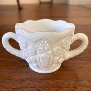 May include: A white, vintage-style sugar bowl with two handles. The bowl has a scalloped rim and a detailed, embossed pattern featuring geometric shapes and a central shield design. The bowl is made of opaque white glass.