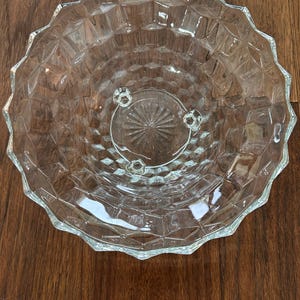 May include: A clear glass serving bowl with a diamond pattern and a scalloped edge. The bowl has three small feet and is sitting on a wooden surface. This vintage bowl is perfect for serving fruit or salads.