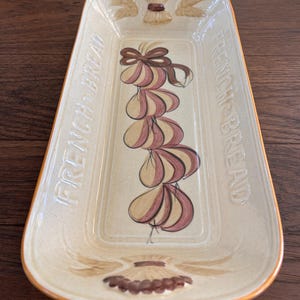 Puede incluir: Plato de cerámica crema para servir pan francés con borde naranja. El plato presenta el texto en relieve "FRENCH BREAD" y un diseño pintado de ajo y trigo. Un artículo de menaje de cocina decorativo.