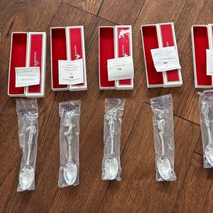 May include: Eight silver-toned metal spoons with decorative handles, each individually sealed in a clear plastic bag. Accompanying the spoons are eight open rectangular boxes with white exteriors, red velvet interiors, and small white paper tags.