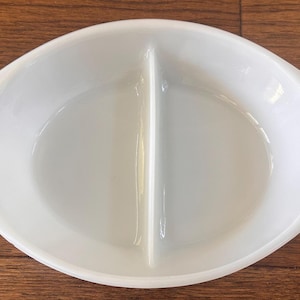 Puede incluir: Plato ovalado blanco dividido con un separador central. El plato tiene lados suavemente curvados y asas integradas. La superficie brillante refleja la luz. Adecuado para servir aperitivos o guarniciones.