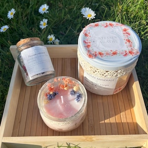 May include: A wooden tray holds a glass jar with a cork lid, a candle in a glass jar, and a container with a floral design lid. The candle is pink with dried flowers. The container has the text "ATELIER AGATHE".