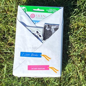May include: A white paper-wrapped package on green grass, with blue and pink tags that read "I Love Books" and "reading DO NOT DISTURB". The package contains a skincare product and stickers.