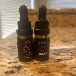 Puede incluir: Dos pequeños frascos cuentagotas de vidrio marrón con tapas negras. Los frascos tienen una etiqueta negra con la palabra "ORYGN" en la parte superior, un diseño de gota roja y el texto "triGLP" debajo. Una botella dice "120 gotas" y la otra "Aprox. 110 gotas".