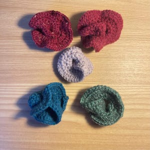 May include: Five handmade crochet items in shades of red, teal, lavender, and green. The items have a ruffled, bunched appearance, and are arranged on a light wood surface. The yarn has a textured appearance.
