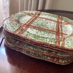 May include: A stack of four square ceramic plates with scalloped edges. The plates feature a white base with a green speckled pattern and a brown plaid design consisting of intersecting lines that cross the surface of each plate.