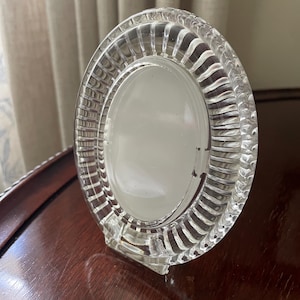May include: A clear glass oval picture frame featuring a decorative border with vertical ribbed detailing. The frame has a smooth, solid center panel and a sturdy base, designed to stand upright on a flat surface for displaying a single photograph.