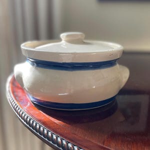 May include: A cream-colored ceramic pot with a matching lid, featuring two side handles and two horizontal blue stripes painted around the body. The pot has a rounded shape and a smooth, glossy finish, presenting a simple and functional design.