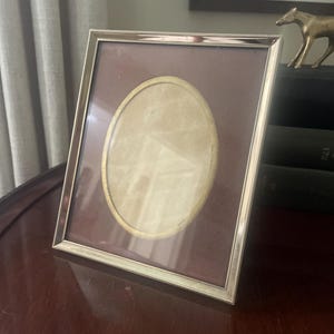 May include: A rectangular gold-toned metal picture frame with a textured rope-style border. The frame features a dark brown matting with a central oval-shaped opening that has a thin gold-colored inner rim, designed to hold a photograph.