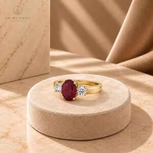 May include: A gold ring featuring a central oval-cut deep red gemstone flanked by two smaller round-cut clear white stones. The band is polished yellow gold, and the setting holds the three stones securely in a classic three-stone arrangement.