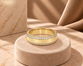 May include: A gold band ring featuring two rows of small, clear, round-cut stones lining the top and bottom edges, with a smooth, polished gold center section. The ring is displayed on a circular, tan-colored, velvet-textured pedestal.