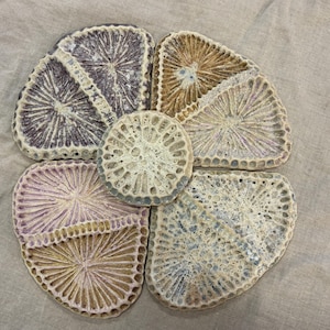 May include: A floral arrangement of handmade coasters in shades of purple, beige, and blue. The coasters are arranged in a flower shape, with a central circular coaster and four petal-shaped coasters surrounding it. The coasters have a textured, porous appearance.