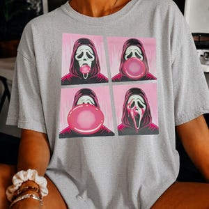May include: A heather grey t-shirt featuring a four-panel graphic print. The design shows a hooded figure with a white mask blowing a pink bubblegum bubble that grows larger in each frame, ending with the bubble popped and covering the mask.
