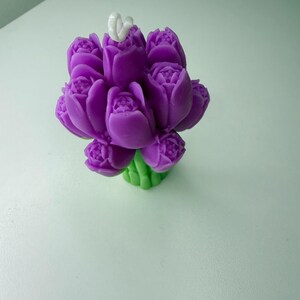 May include: A purple tulip-shaped candle with a white wick. The candle is a bouquet of several tulip blooms, with green stems. The candle is on a white surface.