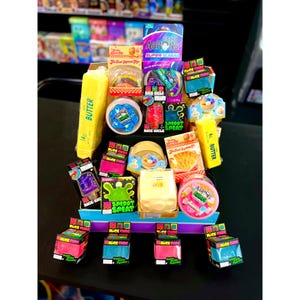 May include: A collection of Crazy Aaron's Thinking Putty and various novelty sensory toys, including butter-shaped containers, popsicle-themed boxes, and colorful slime tubs. The assortment features branded packaging with text like Super Scarab, Nice-Sicle, and Sploot Splat, arranged in a tiered display on a dark surface.