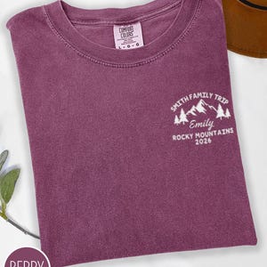 May include: Berry-coloured t-shirt featuring the text "SMITH FAMILY TRIP, Emily, ROCKY MOUNTAINS 2026" and a mountain graphic. The shirt has a crew neck and a tag that says "COMFORT COLORS".