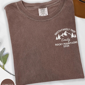 May include: Espresso-coloured t-shirt with a crew neck. The shirt features a white graphic on the upper right chest that reads "Smith Family Trip, Emily, Rocky Mountains 2026" with mountain and tree illustrations. A brown hat is partially visible in the background.