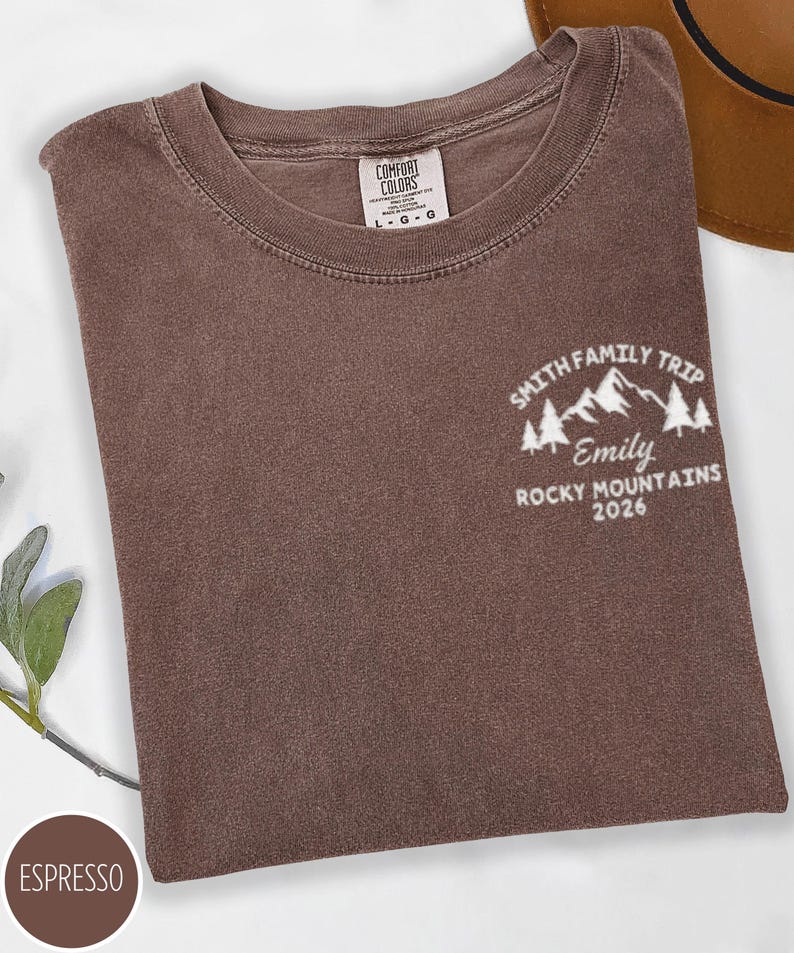 May include: Espresso-coloured t-shirt with a crew neck. The shirt features a white graphic on the upper right chest that reads "Smith Family Trip, Emily, Rocky Mountains 2026" with mountain and tree illustrations. A brown hat is partially visible in the background.