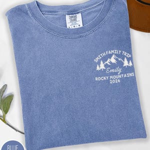 May include: A blue jean coloured t-shirt with a crew neck. The shirt features a white graphic on the front that reads "Smith Family Trip, Emily, Rocky Mountains 2026". The shirt has a Comfort Colors tag.