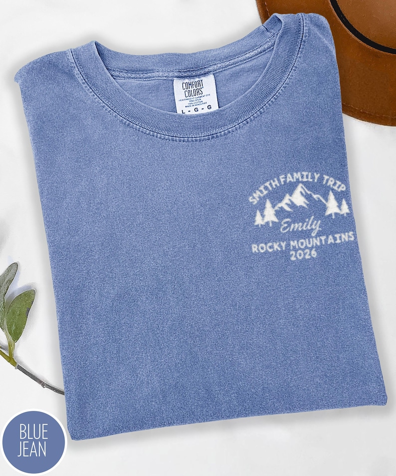 May include: A blue jean coloured t-shirt with a crew neck. The shirt features a white graphic on the front that reads "Smith Family Trip, Emily, Rocky Mountains 2026". The shirt has a Comfort Colors tag.