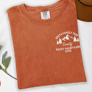 May include: A rust-coloured Comfort Colors t-shirt with a crew neck. The shirt displays a white graphic on the upper left chest, reading "Smith Family Trip, Emily, Rocky Mountains 2026" with mountain and tree illustrations.