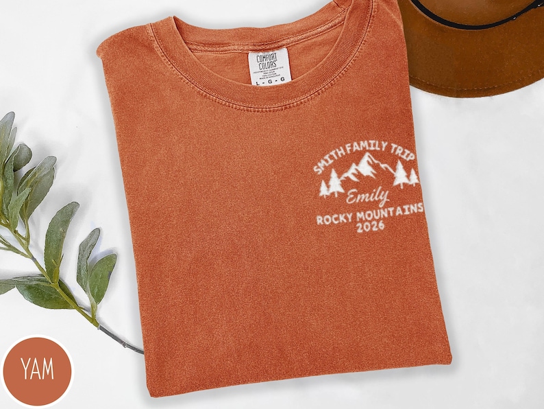 May include: A rust-coloured Comfort Colors t-shirt with a crew neck. The shirt displays a white graphic on the upper left chest, reading "Smith Family Trip, Emily, Rocky Mountains 2026" with mountain and tree illustrations.