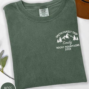 May include: A moss green t-shirt with the text "SMITH FAMILY TRIP Emily ROCKY MOUNTAINS 2026" in white. The shirt has a crew neck and a tag that says "COMFORT COLORS". The shirt is folded.