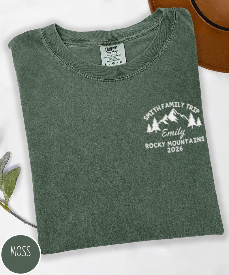 May include: A moss green t-shirt with the text "SMITH FAMILY TRIP Emily ROCKY MOUNTAINS 2026" in white. The shirt has a crew neck and a tag that says "COMFORT COLORS". The shirt is folded.