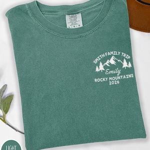May include: A light green t-shirt featuring the text "SMITH FAMILY TRIP, Emily, ROCKY MOUNTAINS 2026" and a mountain graphic. The shirt has a crew neck and a "COMFORT COLORS" tag.