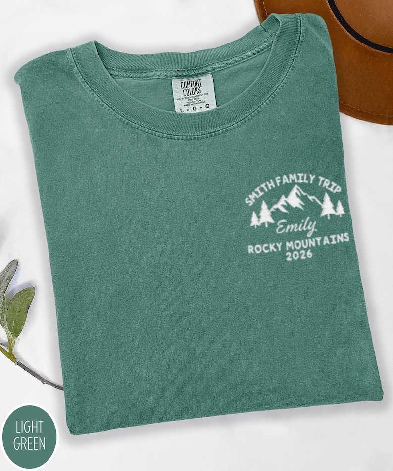May include: A light green t-shirt featuring the text "SMITH FAMILY TRIP, Emily, ROCKY MOUNTAINS 2026" and a mountain graphic. The shirt has a crew neck and a "COMFORT COLORS" tag.