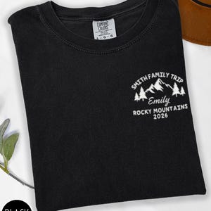 May include: Black t-shirt with a white graphic of mountains, trees, and the text "SMITH FAMILY TRIP, Emily, ROCKY MOUNTAINS 2026." The shirt has a crew neck and a tag that reads "COMFORT COLORS." A black circle with the word "BLACK" is in the lower left corner.