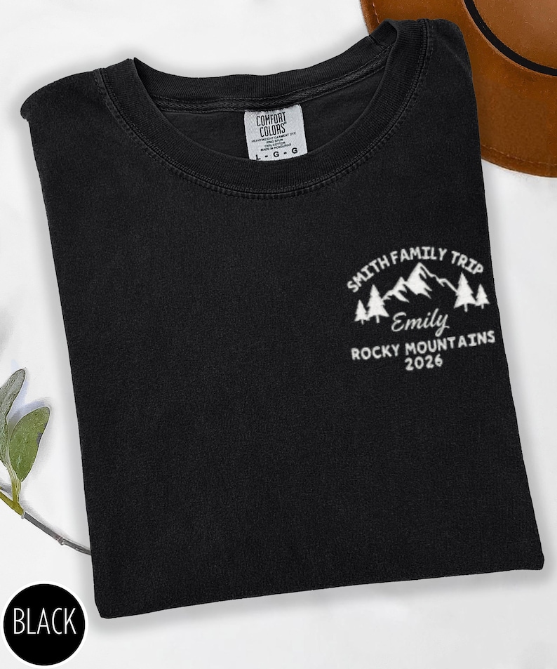 May include: Black t-shirt with a white graphic of mountains, trees, and the text "SMITH FAMILY TRIP, Emily, ROCKY MOUNTAINS 2026." The shirt has a crew neck and a tag that reads "COMFORT COLORS." A black circle with the word "BLACK" is in the lower left corner.