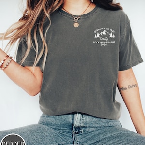 May include: Dark grey t-shirt with a white graphic on the upper left chest. The graphic includes text that reads "SMITH FAMILY TRIP", "Emily", "ROCKY MOUNTAINS 2026", and a mountain and tree design. The shirt is paired with blue jeans.