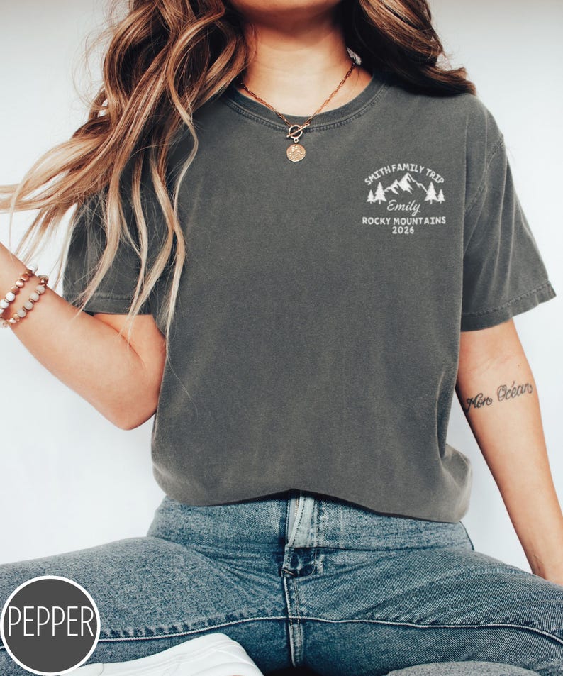 May include: Dark grey t-shirt with a white graphic on the upper left chest. The graphic includes text that reads "SMITH FAMILY TRIP", "Emily", "ROCKY MOUNTAINS 2026", and a mountain and tree design. The shirt is paired with blue jeans.
