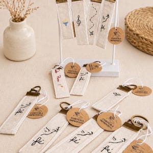 Puede incluir: Una colección de llaveros de tela color hueso con varios diseños bordados en negro, incluyendo la palabra fé, notas musicales y un sol. Cada llavero tiene una argolla de metal color bronce y una etiqueta redonda de papel marrón Costura Improvável.