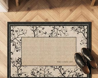 May include: Rectangular doormat with a black border and a beige center. The mat features a tree branch design in black around the edges and the words "THE GOOD LIFE" printed in the lower right corner. Two black leather shoes are placed on the mat.