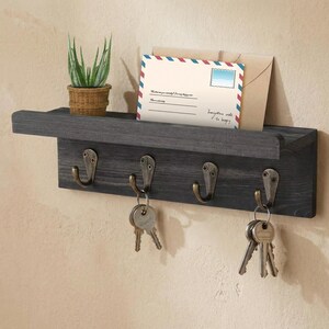 May include: A gray wooden key holder with four metal hooks. A small potted plant and an envelope with text are on the shelf. Keys hang from the hooks. The text on the envelope reads "Everything adds to happy."