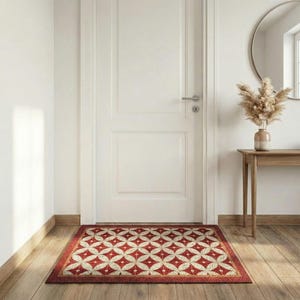 May include: A rectangular doormat with a geometric pattern of red and cream-colored interlocking circles. The mat has a red border and is placed in front of a white door on a wooden floor. The scene includes a round mirror and a small table with a vase.