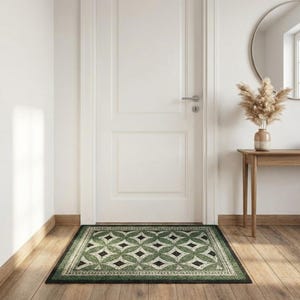 May include: A rectangular green and black patterned doormat with a geometric design is placed in front of a white door. The mat features a repeating pattern of diamonds and leaf-like shapes. The floor is made of wood.