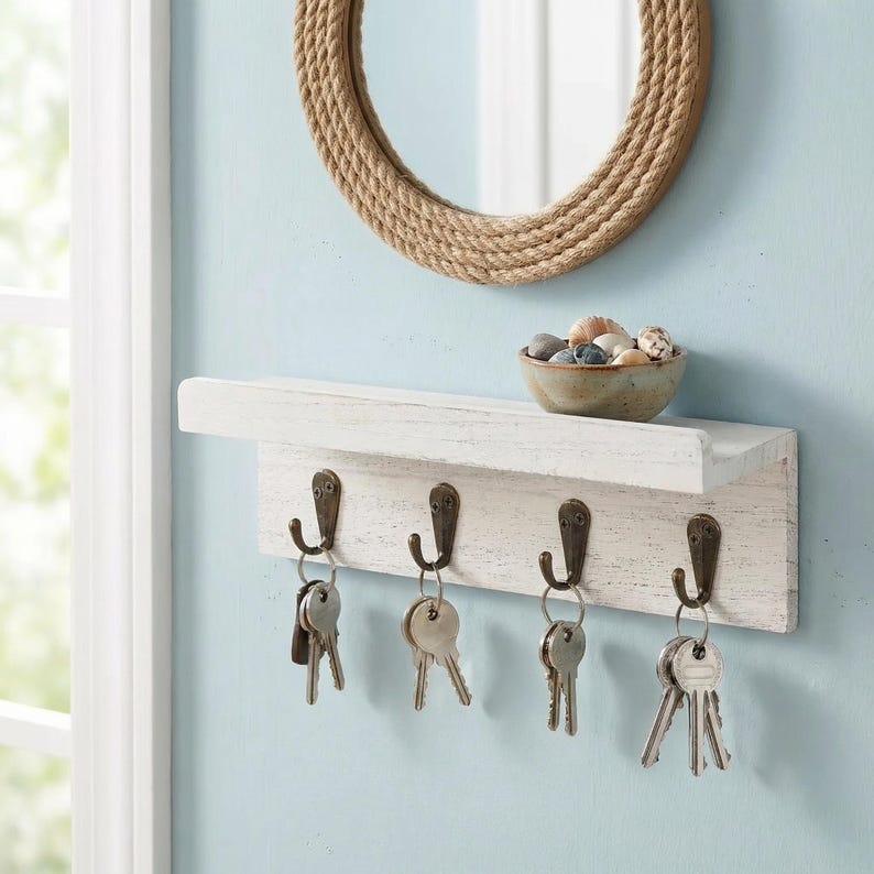 May include: A white wooden key holder with four metal hooks, holding various keys. Above the hooks is a small shelf with a ceramic bowl filled with stones. A round mirror with a rope-like frame hangs above the shelf.