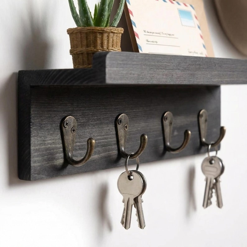 May include: A dark gray wooden key holder with four antique brass hooks. The key holder has a shelf above it, with a small potted plant and a decorative envelope. Two sets of keys hang from the hooks.